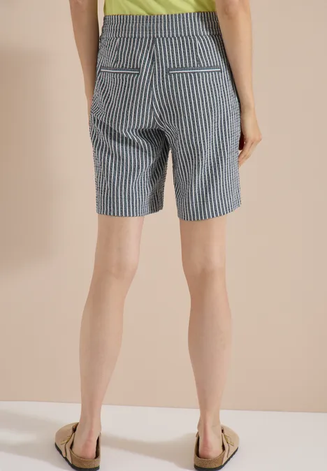 Rückansicht einer Person in blau-weiß gestreiften Shorts und Sandalen vor beigem Hintergrund.