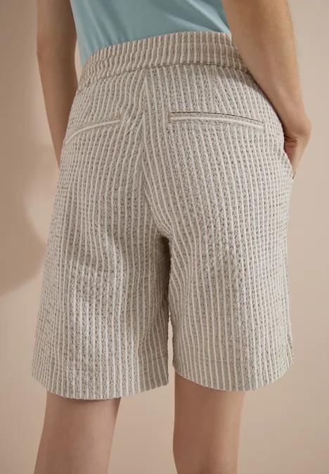Rückansicht einer Person in beige-weiß gestreiften Bermudashorts mit Taschen und hellblauem Oberteil vor beige Hintergrund.