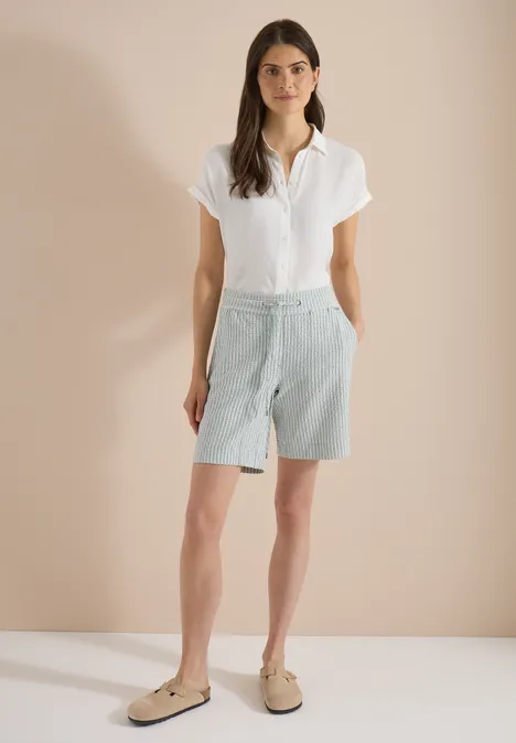 Frau in weißem Kurzarmhemd, hellblauen Bermudashorts und beigen Loafers vor neutralem Hintergrund.
