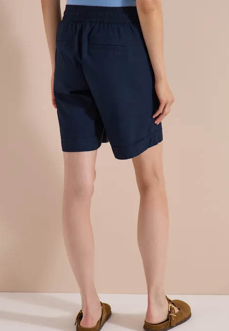 Rückansicht einer Person in marineblauen Bermudashorts und braunen Mokassins vor hellem Hintergrund.