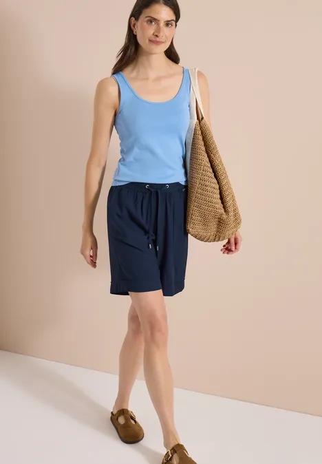 Frau in blauem Tanktop, dunkelblauen Shorts und braunen Sandalen vor beigem Hintergrund mit großer Gewebetasche.