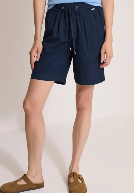 Eine Person trägt dunkelblaue, knielange Shorts mit Kordelzug und Taschen, dazu Sandalen.