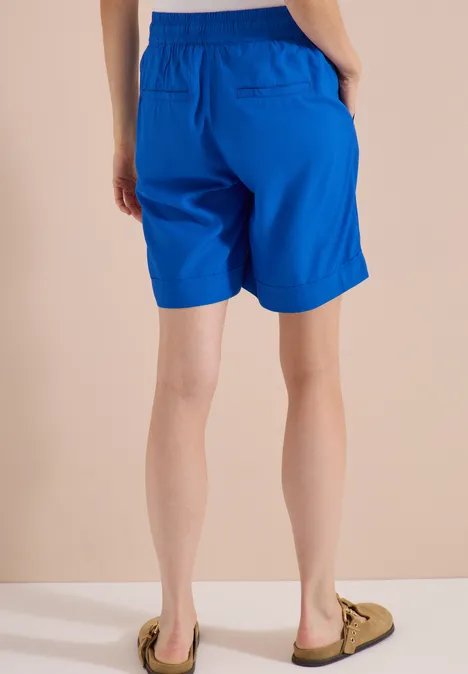 Eine Person von hinten in knielangen, leuchtend blauen Shorts und hellen Slippern vor einem neutralen Hintergrund.