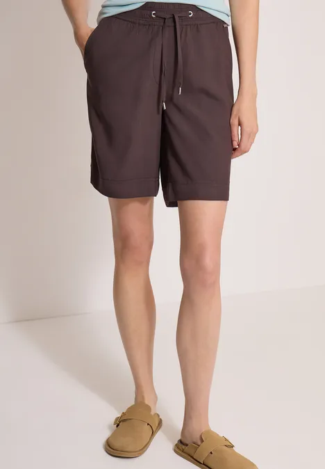 Eine Person trägt dunkelbraune Shorts mit Kordelzug und beigefarbene Slip-on-Sandalen.