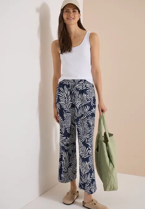 Pantalon à jambes larges avec imprimé et galons universal blue Femme en débardeur blanc et pantalon ample bleu et blanc à motif de palmiers, sac vert et chaussures beiges.