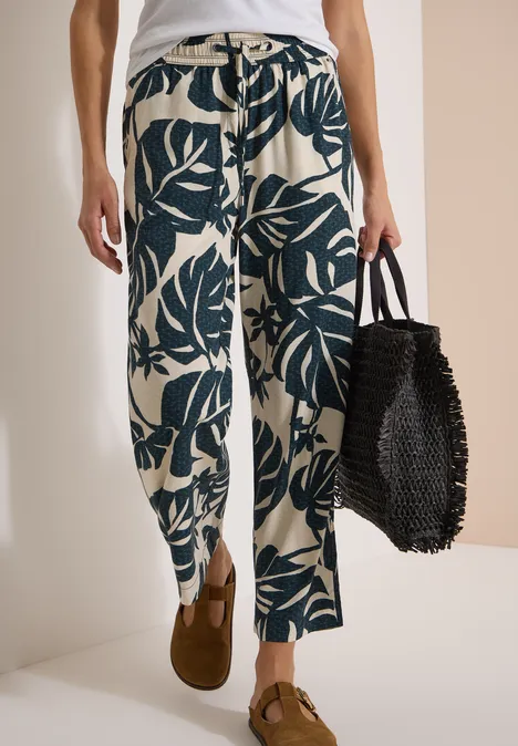 Weite, helle Hose mit dunkelgrünem Blattmuster, braune Loafers und schwarze, geflochtene Tasche.
