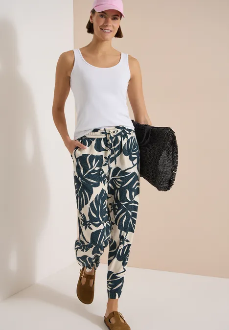 Fröhliche Frau im weißen Top, gemusterter Hose und rosa Cap, mit schwarzer Strandtasche.
