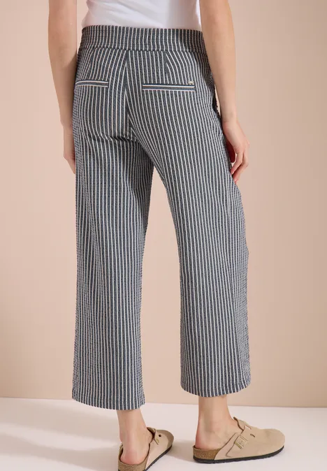 Vrouw van achteren in knielange, zwart-wit gestreepte broek en beige loafers voor een beige achtergrond.