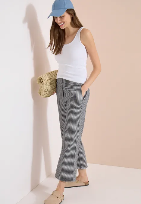 Vrouw in witte top, grijze geruite broek, blauwe pet en stro tas leunend tegen een muur.