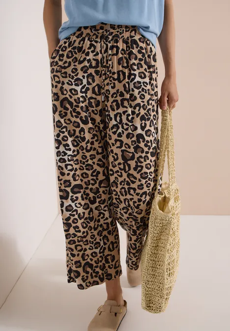 Luchtige, wijdvallende broek met luipaardprint, gecombineerd met sandalen en een gehaakte rieten tas.