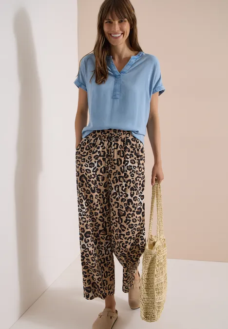 Vrouw met lichtblauw poloshirt, wijde luipaardbroek en strooien tas voor een neutrale achtergrond.