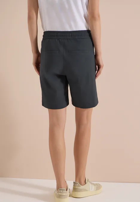 Achteraanzicht van een persoon in een wit topje, zwarte short en beige sneakers voor een lichte achtergrond.