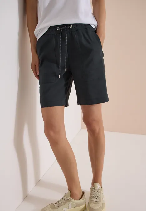 Persoon in zwarte shorts met trekkoord, witte top en lichte sneakers tegen een minimalistische achtergrond.