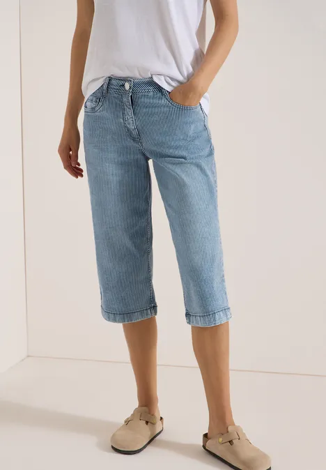 Person steht vor hellem Hintergrund in weißem Shirt, hellblauen Capri-Jeans und beigen Slippern.