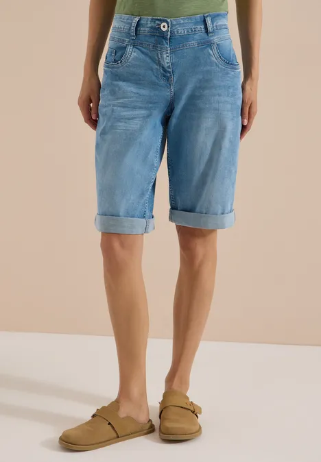 Person trägt hellblaue Jeans-Bermudashorts und beigefarbene Sandalen vor neutralem Hintergrund.