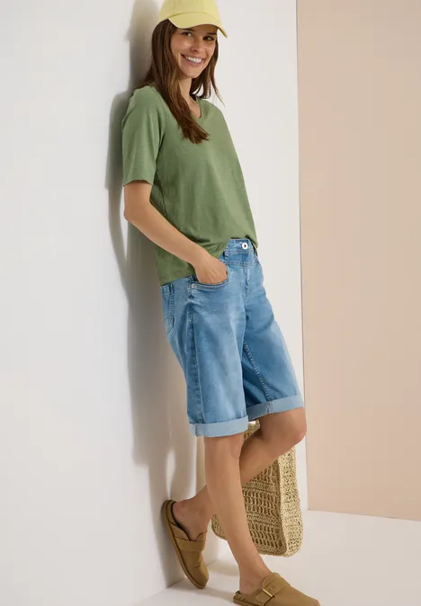 Junge Frau lehnt entspannt an der Wand, trägt grünes T-Shirt und knielange Jeans-Shorts.