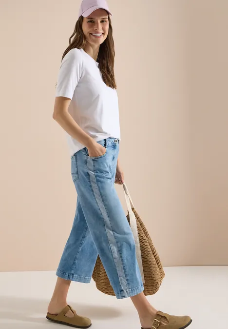 Frau in weißem T-Shirt, weitem Jeans-Culotte, Basttasche und flachen Sandalen vor neutralem Hintergrund.