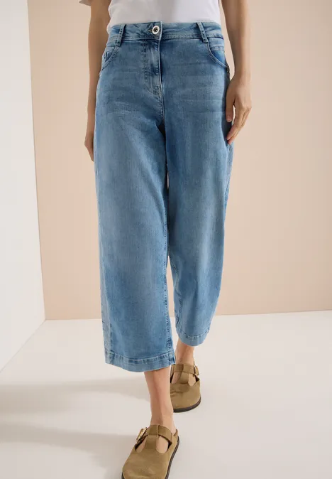 Frau in weiten, hellblauen Cropped-Jeans bis zur Wade und braunen Riemchen-Schuhen vor schlichtem Hintergrund.