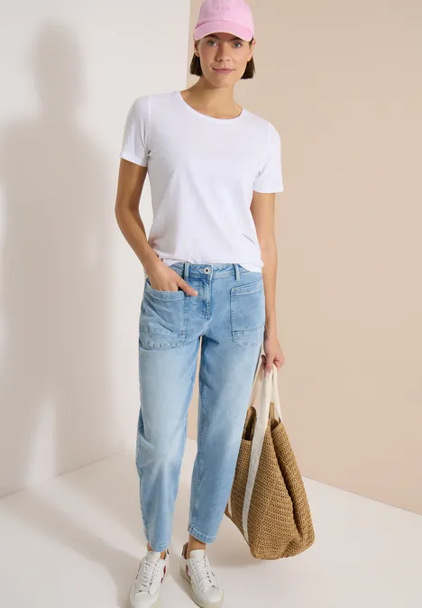 Frau in weißem T-Shirt, hellblauen Jeans und weißen Sneakern hält eine große Strohtasche.