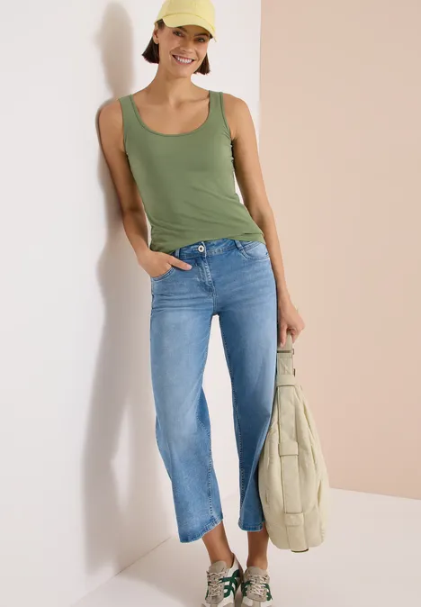 Eine Person in grünem Top und blauen Jeans lehnt an der Wand und hält eine helle Tasche.