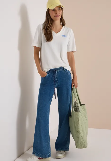 Eine Frau trägt ein weißes V-Neck-Shirt, blaue Schlagjeans, eine hellgelbe Cap und hält eine olivgrüne Tasche.