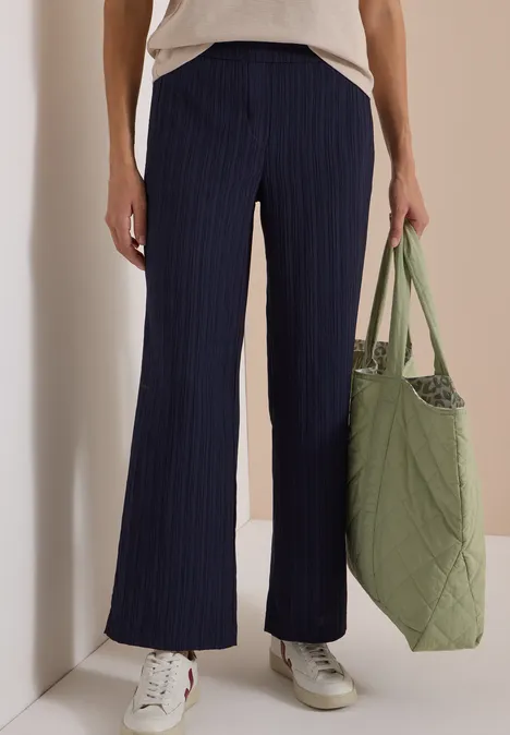 Frau in locker sitzender navy Nadelstreifenhose, weißem Shirt, weißen Sneakern und grüner, gesteppter Tasche.