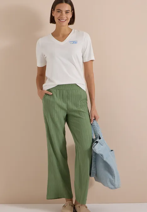 Frau trägt weißes V-Neck-Shirt, weite grüne Hose und gestreifte Tasche vor neutralem Hintergrund.