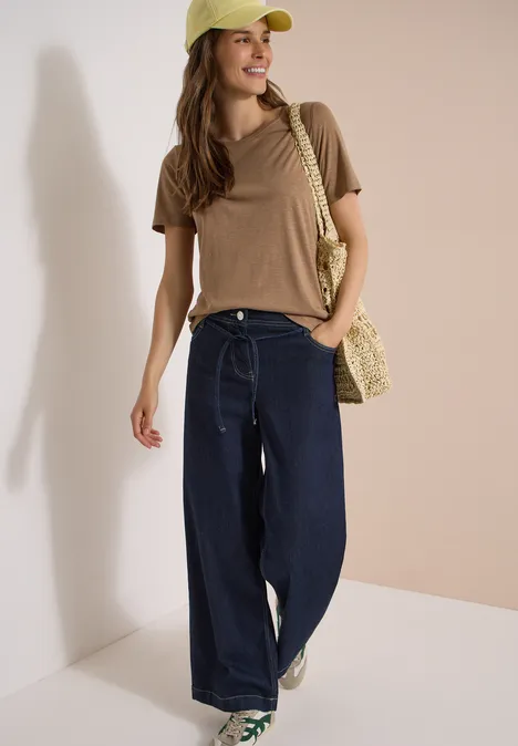 High Waist Wide Leg Jeans im Loose Fit rinsed wash Frau in braunem T-Shirt, weiten Jeans, grünen Sneakern, Strohtasche und Cap vor neutralem Hintergrund.