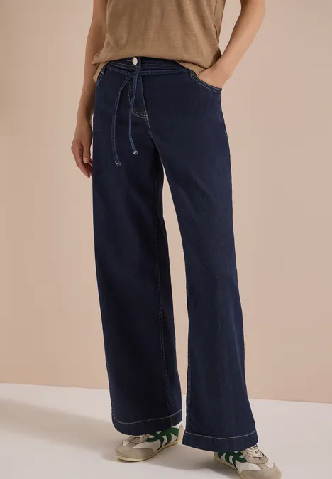 High Waist Wide Leg Jeans im Loose Fit rinsed wash Frau trägt weite, dunkelblaue Jeans mit Kordelzug und Sneakers vor hellem Hintergrund.