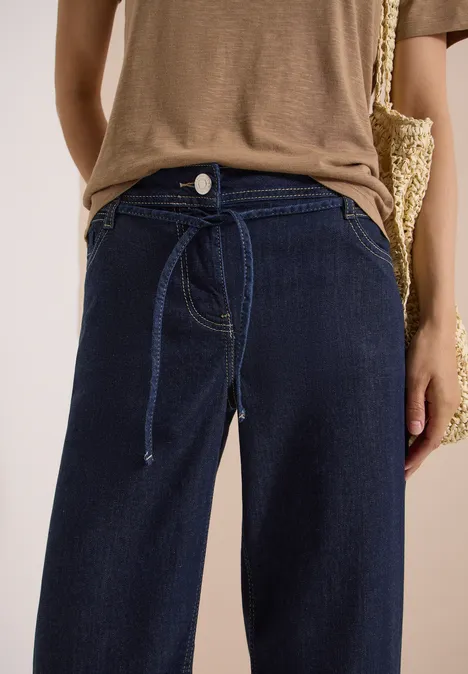 High Waist Wide Leg Jeans im Loose Fit rinsed wash Person trägt hoch geschnittene, dunkelblaue Jeans mit Bindegürtel, kombiniert mit braunem Oberteil und Basttasche.
