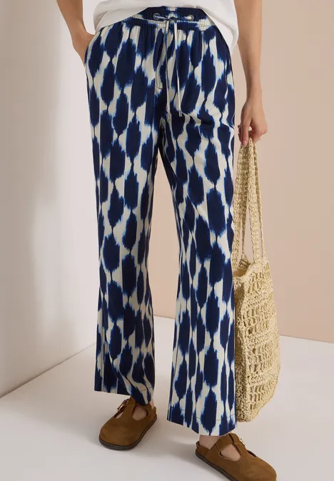 Weite Hose mit blau-weißem Ikat-Muster, weißes Oberteil, Basttasche und braune Mokassins.