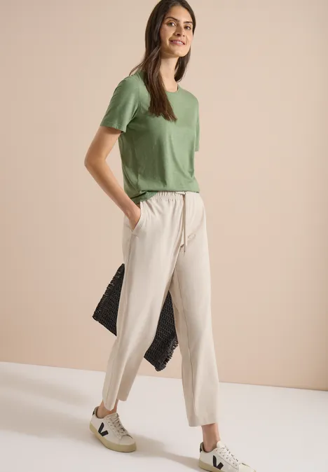 Broek met High Waist en Wide Leg pijpen in een Loose Fit pasvorm cashmere beige melange Vrouw in groen T-shirt, beige broek en witte sneakers staat zijwaarts voor een beige achtergrond.