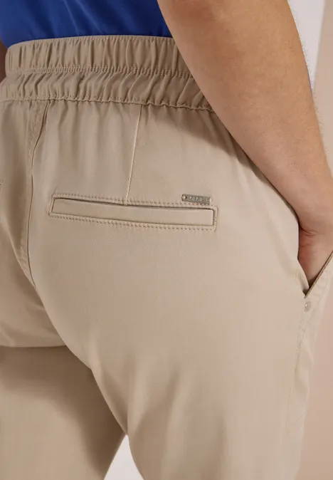 Beige Hose mit elastischem Bund, Gesäßtasche mit Markenlabel, eine Hand steckt in der vorderen Hosentasche.