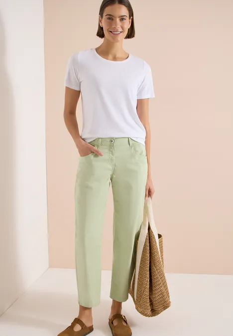 Spodnie z szerokimi nogawkami z tkaniny o fakturze papieru raw matcha lime Kobieta w białym t-shircie i pastelowych zielonych spodniach, trzyma dużą plecioną torbę na jasnym tle.