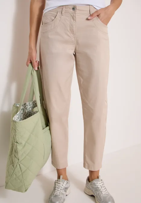 Spodnie z szerokimi nogawkami z tkaniny o fakturze papieru cashmere beige Osoba nosi beżowe chino, białe trampki i trzyma zieloną, pikowaną torbę.