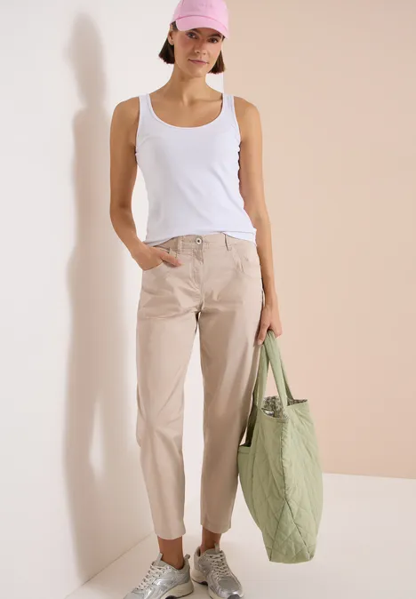 Spodnie z szerokimi nogawkami z tkaniny o fakturze papieru cashmere beige Kobieta w białym topie, beżowych spodniach i białych trampkach trzyma dużą jasnozieloną torbę.
