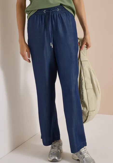 Person trägt weite, dunkelblaue Stoffhose mit Tunnelzug, silberne Sneaker und eine leichte Umhängetasche.