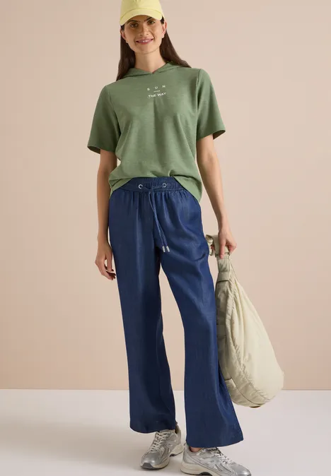 Junge Frau in grünem T-Shirt, weitem blauen Hosen und Cap, lässig stehend mit großer Tasche.