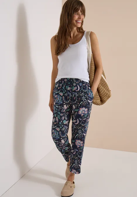 Frau in weißem Top, gemusterten Paisley-Hosen, Basttasche und beigen Slippern vor neutraler Wand