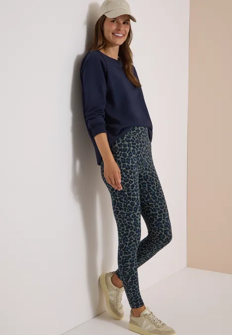 Legging met luipaardprint dried olive green Vrouw leunt tegen muur, draagt pet, donkerblauwe trui, patroonlegging en lichte sneakers.