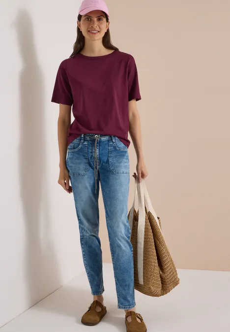 Eine Frau in rosa Cap, burgunderrotem T-Shirt, blauen Jeans und mit einer beigefarbigen Tasche vor hellem Hintergrund.