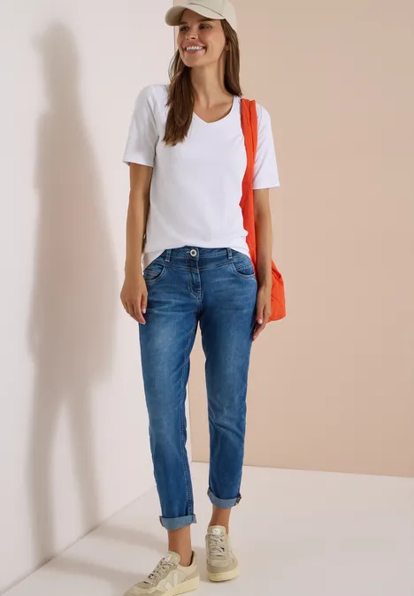 Frau in weißem T-Shirt, Jeans, Sneakers und Cap, mit orangem Schulterbeutel, steht lächelnd vor hellem Hintergrund.