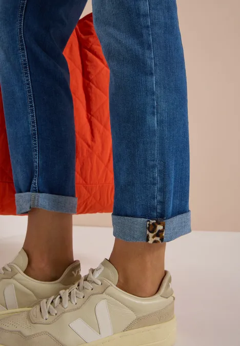 Hochgekrempelte Jeans, helle Sneaker und roter Steppmantel im Hintergrund.