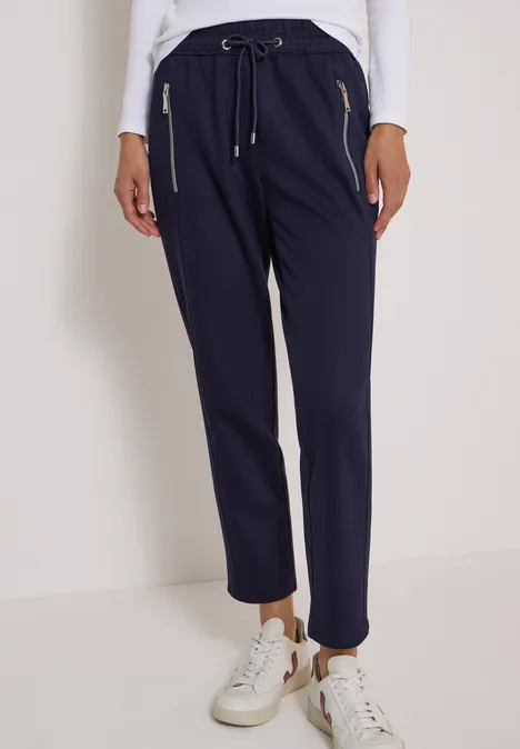 Pantalon taille mi-haute, coupe Slim Leg et style décontracté urban dark blue Dunkelblaue Damenhose mit elastischem Bund, Kordelzug und goldenen Reißverschlusstaschen, weiße Sneaker.