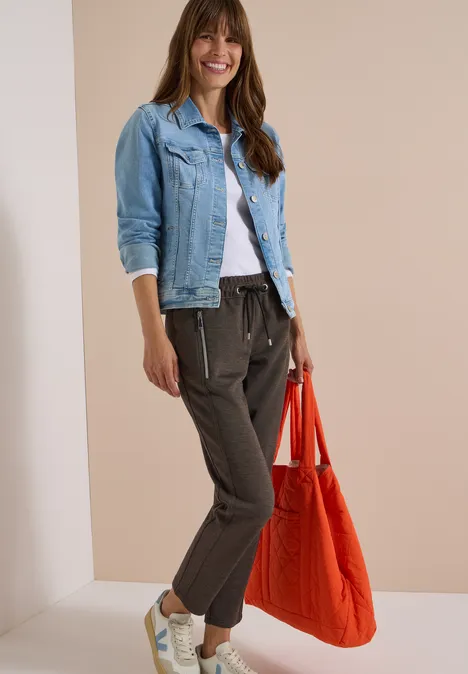 Pantalon taille mi-haute, coupe Slim Leg et style décontracté creamy mocha Fröhliche Frau im weißen T-Shirt, Jeansjacke, dunkelgrauen Jogginghosen, weißen Sneakers und roter Tasche.