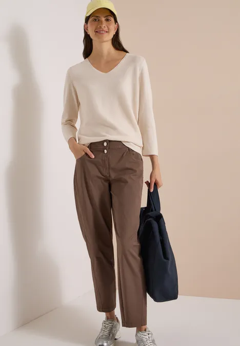 Eine Frau in einem hellen V-Pullover, braunen Hosen und Cap, trägt eine dunkelblaue Tasche.