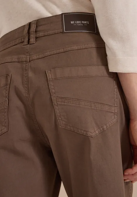 Rückansicht einer Person in braunen Hosen mit Gesäßtaschen und Label am Bund, dazu helle Oberbekleidung.