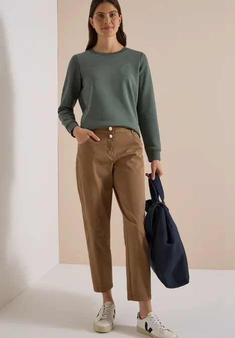 Mid Waist Barrel Leg Hose im Casual Fit creamy mocha Frau im grünen Pullover, beigen Hosen und weißen Sneakern hält blaue Tasche in hellem Raum.