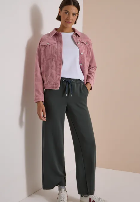 Frau vor neutralem Hintergrund mit rosa Jeansjacke, weißem Shirt, weiten schwarzen Hosen und weißen Sneakern.