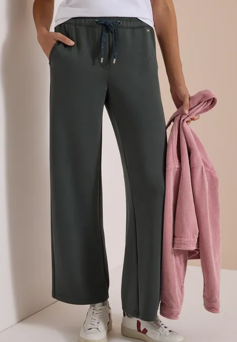 Dame trägt weite, dunkelgraue Jogginghose mit Tunnelzug, weiße Sneaker und hält ein rosa Sweatshirt.